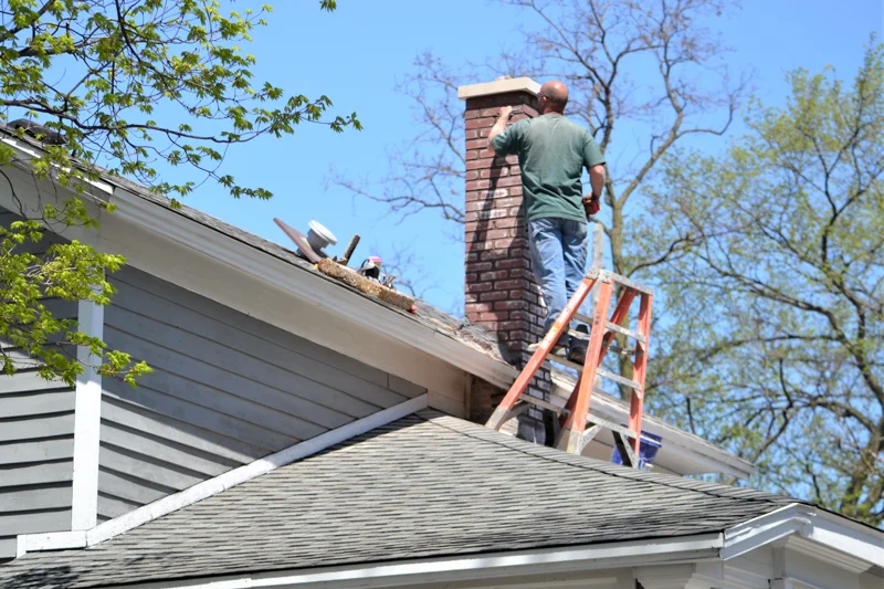 Worker on ladder repairing brick chimney on sloped roof performing chimney repair